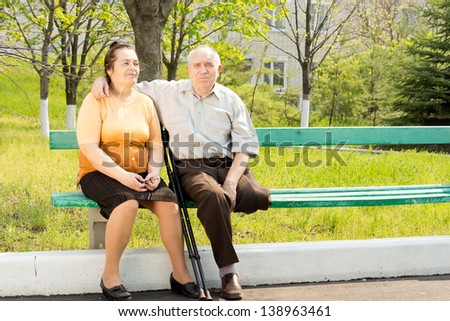 Elderly couple sitting close together on a park bench enjoying the sunshine - the husband has one leg amputated and is using crutches