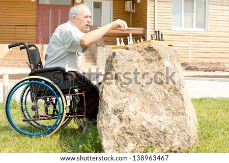 Elderly handicapped man playing chess sitting in his wheelchair in front of his house in the garden with the chessboard on top of a convenient rock