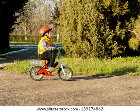 Cute little boy learning to ride pedalling his bicycle along a rural country lane dressed in his safety helmet and bright orange high visibility jacket