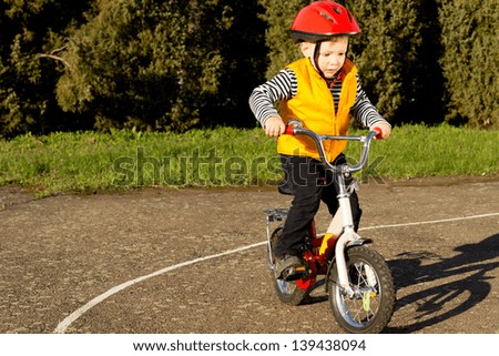 Cute young boy dressed in a colourful red safety helmet and orange high visibility jacket practising riding his bike on a quiet country lane