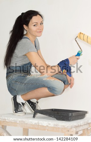 Smiling woman painting the wall of her home balanced on a wooden trestle with a paint tray at her feet and roller in her hand