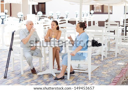 Elderly amputee on crutches enjoying a drink with his family at an open-air restaurant sitting with his wife and daughter at a shady table