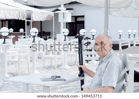 Elderly handicapped man sitting at a table at an outdoor restaurant with his crutches in his hand looking at the camera with a serious expression