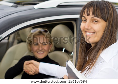 Smiling saleslady congratulating the new owner of a car as they shake hands to seal the deal through the open window of the vehicle