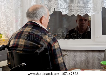 Elderly man in a wheelchair sitting waiting at a window with his back to the camera staring through the glass into the dark night