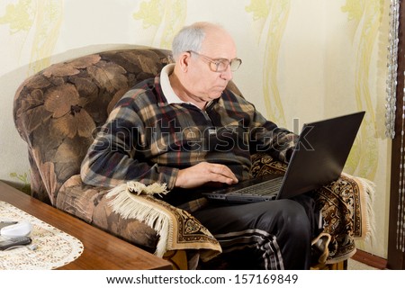 Elderly man sitting in a comfortable armchair at home with his computer on his lap surfing the internet on a laptop