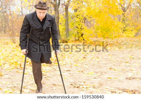 Elderly disabled gentleman with one leg amputated walking on crutches in an autumn park with colourful yellow foliage