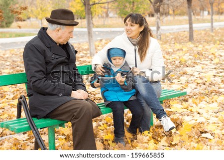 Three generations - a disabled grandfather on crutches, an attractive smiling mother and her cute young son who is playing on a tablet-pc, all sitting together on a wooden park bench in autumn