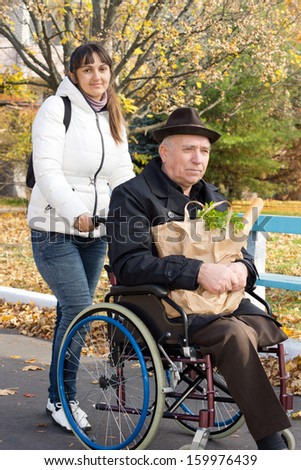 Smiling woman helping her disabled elderly father taking him out grocery shopping pushing him along the street in his wheelchair
