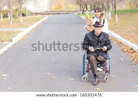 Female carer helping a disabled elderly man in a wheelchair pushing him along the street and carrying a bag of groceries from the supermarket