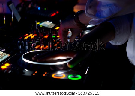 Colourful DJ music deck at night with all the controls illuminated and the hand of the disc jockey suspended above the vinyl record on the turntable to mix and scratch the audio for the disco