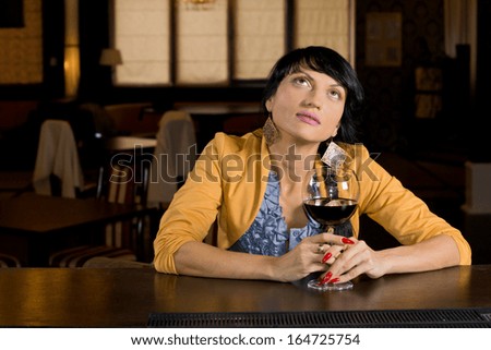 Thoughtful woman sitting drinking red wine at the bar staring up into the air as she reminisces or daydreams