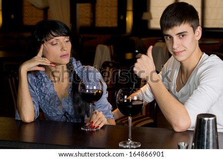 Handsome young man drinking a glass of red wine at a bar with a female friend giving a thumbs up of approval to show that he is enjoying himself and all is going well