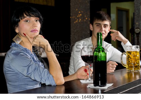 Sophisticated young woman drinking at a bar enjoying a bottle of red wine turning to look at the camera with a man in the background