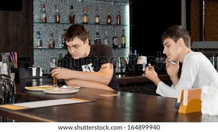 Two men friends enjoying themselves having a drink of beer at the bar with one busy having a conversation on his mobile phone