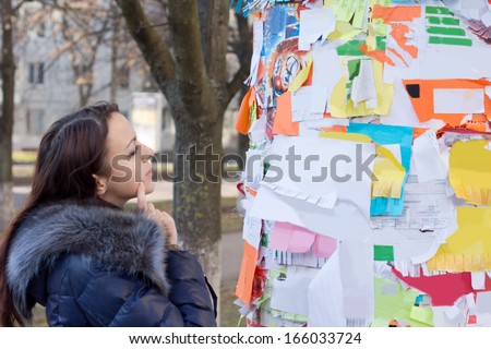 Pensive woman reading advertisements on a public notice board in a town square or park looking at them thoughtfully as she makes a decision