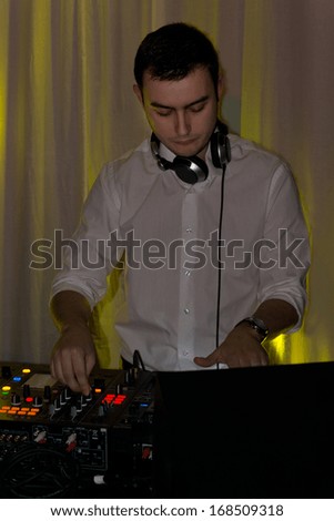 Handsome disc jockey mixing music standing at his deck at a party or disco dance with his earphones around his neck concentrating on his deck