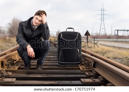 Bored young man waiting for the arrival of a train squatting on the sleepers in the centre of the railway line with his head on his hand and his suitcase alongside