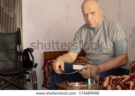 Senior handicapped man taking his blood pressure as he sits at home alongside his wheelchair using a pressure cuff and sphygmomanometer