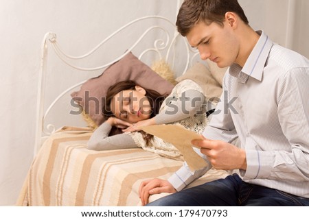 Stylish young man in the bedroom reading a letter while sitting on the edge of a vintage wrought iron bed watched by his wife or girlfriend