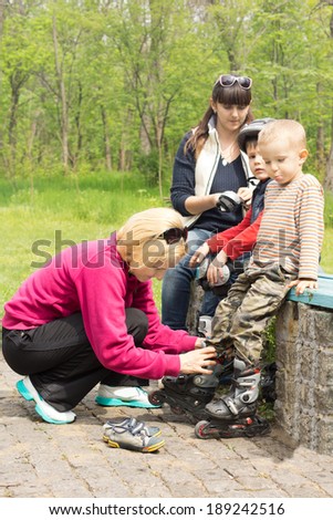 Two mothers sitting on a stone wall fitting their young sons with roller blades, knee pads and helmets as they prepare to go roller skating