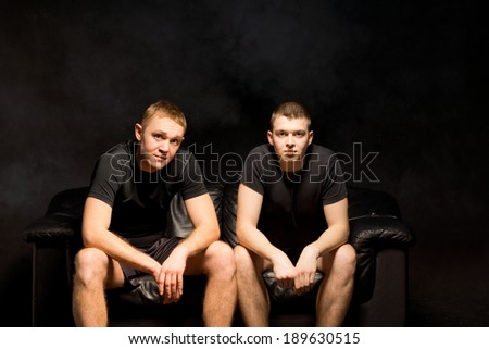 Two serious fit young men sitting side by side on a black sofa in black shorts and t-shirts looking thoughtfully at the camera on a dark background with copyspace