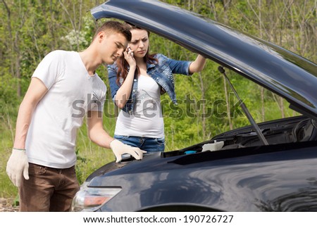 Young mechanic looking at a car engine after a call-out from an attractive young woman for a roadside breakdown as she stands behind him chatting on her mobile phone