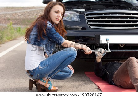 Attractive young woman kneeling in front of her broken down car handing a spanner to a mechanic working underneath the engine compartment