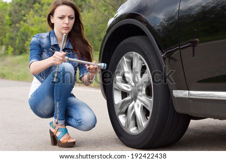 Confused woman looking at a wheel spanner with a puzzled expression as she kneels in the road alongside her car which has broken down