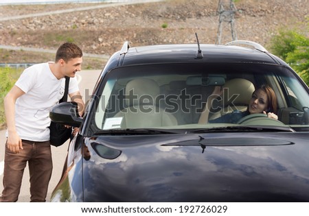 Young male hitchhiker looking for a lift talking to a female driver through the passenger window of her car as he tries to persuade her to give him a ride