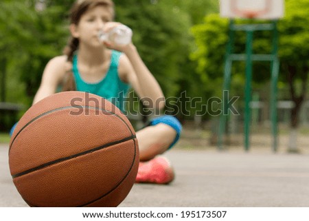 Young basketball player drinking water as she sits cross-legged on the court with the ball in front of her, focus to the ball