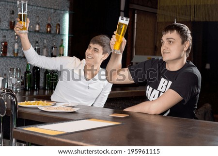 Two young men sitting at the bar counter in a club raising their beer glasses high in the air in a toast as they celebrate together
