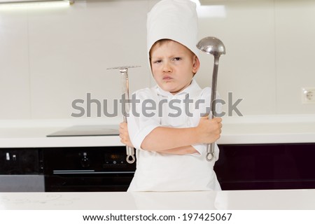 Young boy chef protecting his secret recipe standing gripping his utensils with folded arms and a pugnacious expression
