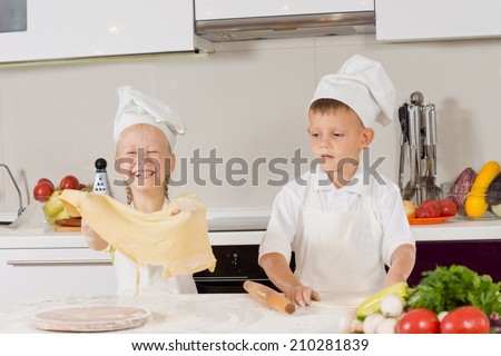 Two young children having fun making pizza with the little girl giggling as she holds aloft a large sheet of pastry that the little boy has just finished rolling out