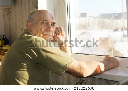 Lonely senior man sitting at a window turning to look at the camera with a serious thoughtful expression
