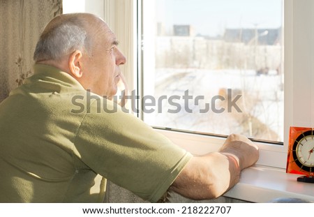 Senior man standing reminiscing as he stares out of a window with a faraway expression as he recalls nostalgic old memories