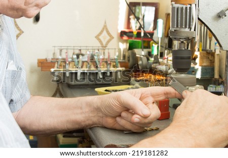 Elderly man using a small electric bench grinder to sharpen a tool or grind down a metal bar as he sits at his workbench