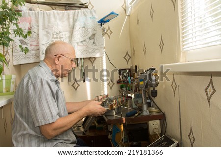 Retired man working at home on his handicrafts sitting at a compact workbench in his house using precision tools and small scale electrical equipment and power tools