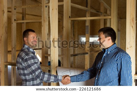 Two men shaking hands in a half constructed timber frame house with a building blueprint alongside them as they signal their satisfaction with the progress