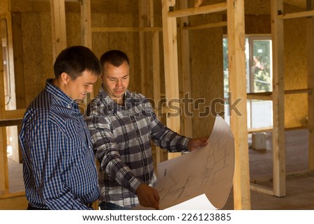 Two builders inside a half completed timber frame house standing having a discussion over a blueprint of the building