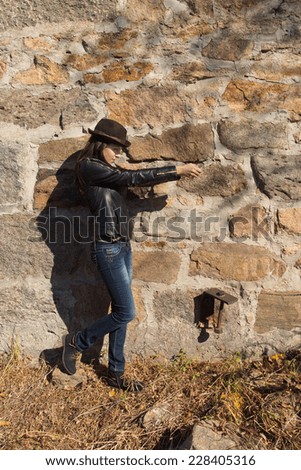 Slender chic young woman standing sideways in front of a stone wall in her leather jacket and trendy hat in the sunshine, copyspace above