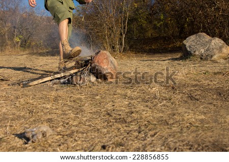 Young man stamping out the smouldering embers a fire in a campsite with his booted foot to avoid the danger of it spreading, with copyspace, low angle view of his legs