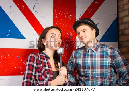 Close up Young Woman with Microphone, Showing Wide Open Eyes Expression, Interviewing Young Male Guest in Trendy Outfit in Front Huge British Flag Print.
