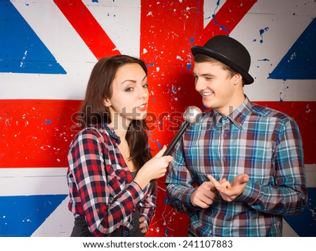 Female British chat show host interviewing a handsome young man in a bowler hat in front of a Union Jack flag painted on the wall
