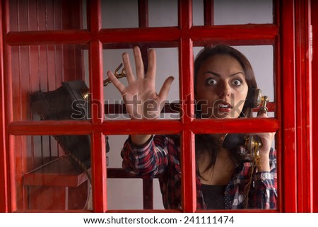 Terrified woman trapped in a telephone booth staring out with a wild wide eyed expression of panic as she holds the vintage handset in her hand