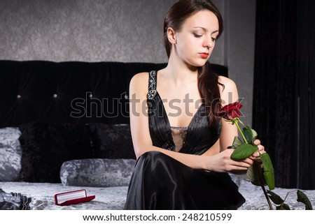 Gorgeous Young Woman in Black Dress Sitting at the Bedroom While Holding Red Rose Flower.