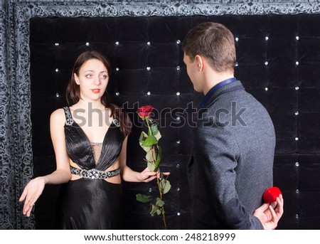 Close up Young Man with Jewelry Box Behind his Back Offering a Rose Flower to Unhappy Lady in Sexy Black Evening Gown.