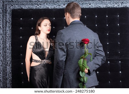 Close up Young Man Talking to a Pretty Lady, in Sexy Black Dress, with Red Rose Flower on his Back. Captured on Sparkling Black Background.