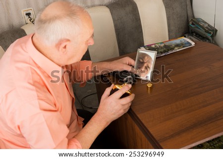 Close up Sitting Old Bald Man with Aftershave Bottle on his Hand Looking at the Small Mirror on Wooden Table.