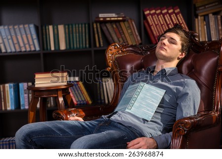 Attractive tired young man asleep in a library reclining in a comfortable armchair with his open book resting on his chest